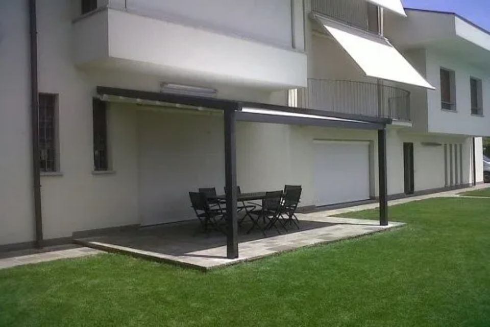 Patio con tavolo e sedie sotto un pergolato scuro, accanto a un edificio bianco e all'erba verde.