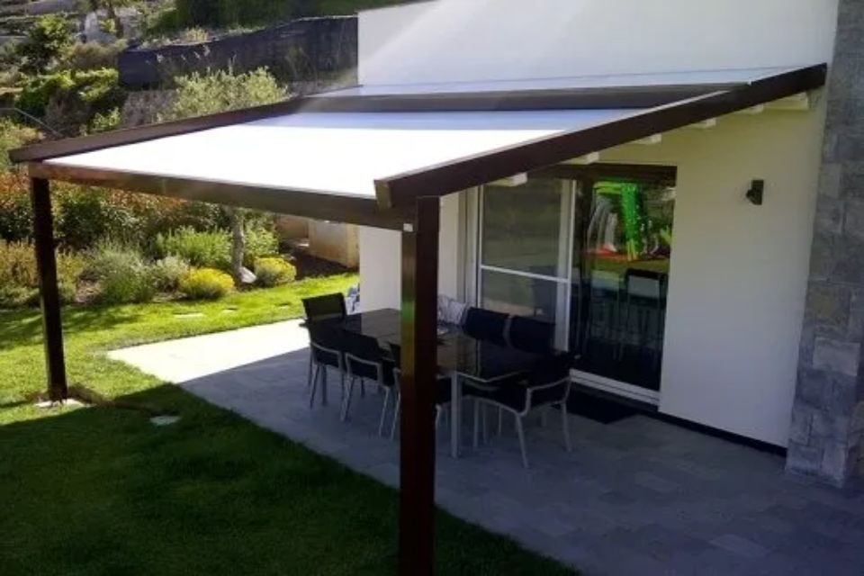 Patio con pergolato marrone e tenda da sole bianca, sopra un tavolo e delle sedie accanto a una casa con un prato verde.