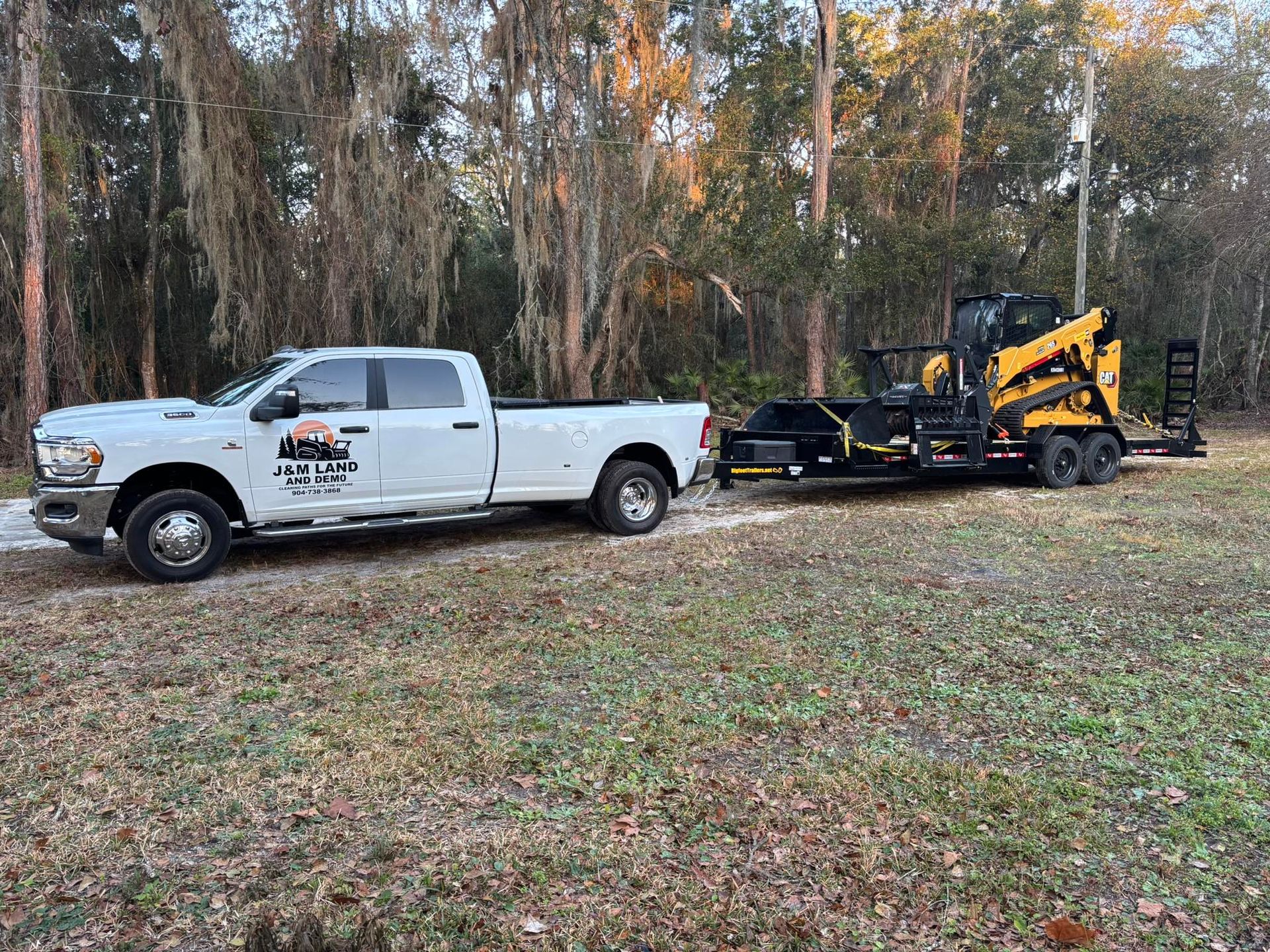White pickup truck towing a black-and-yellow skid steer on a trailer in a wooded field