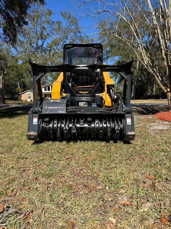 Front view of a yellow skid-steer loader with a black brush attachment on grass under trees