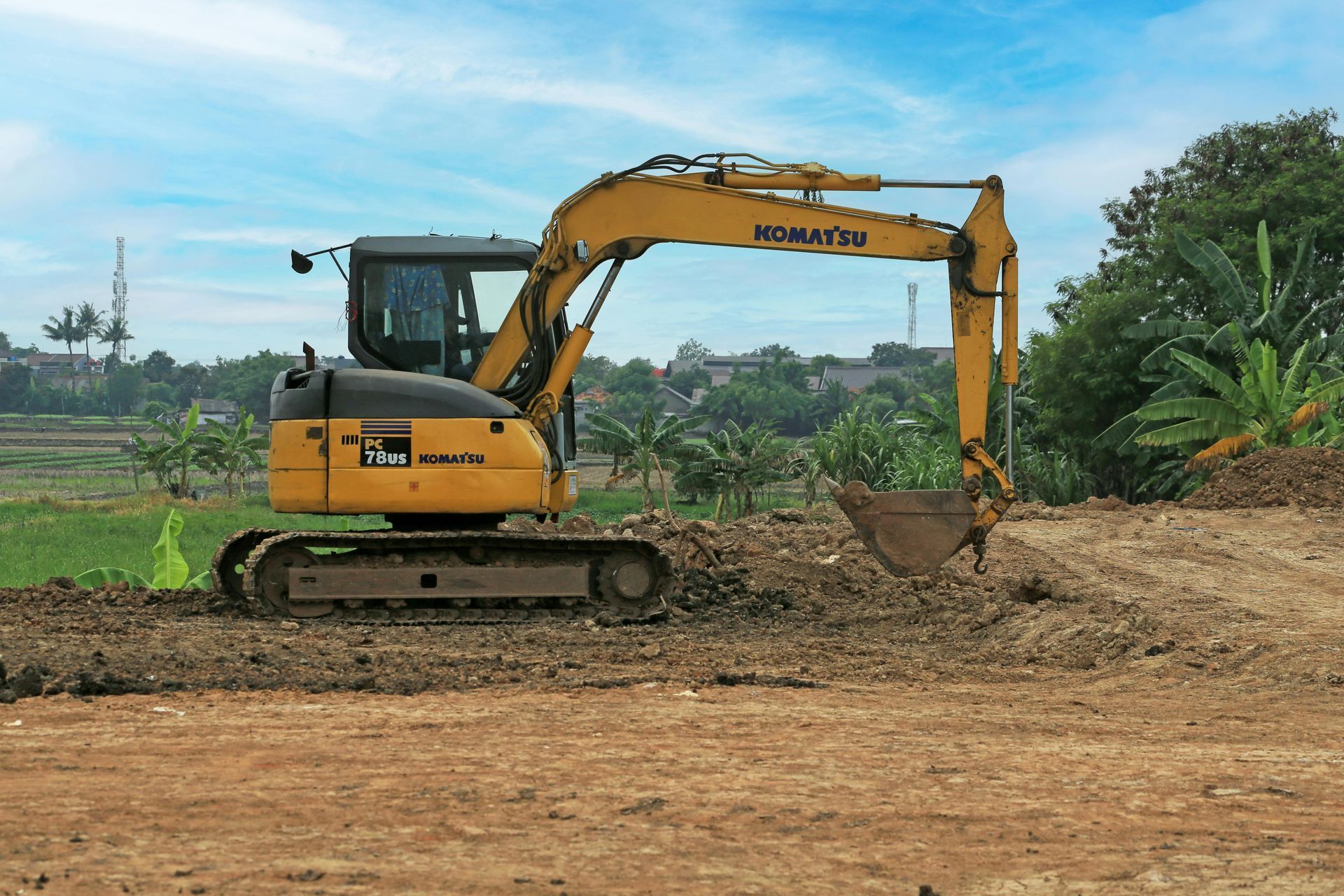 Yellow excavator on a dirt field, digging near green trees under a cloudy sky