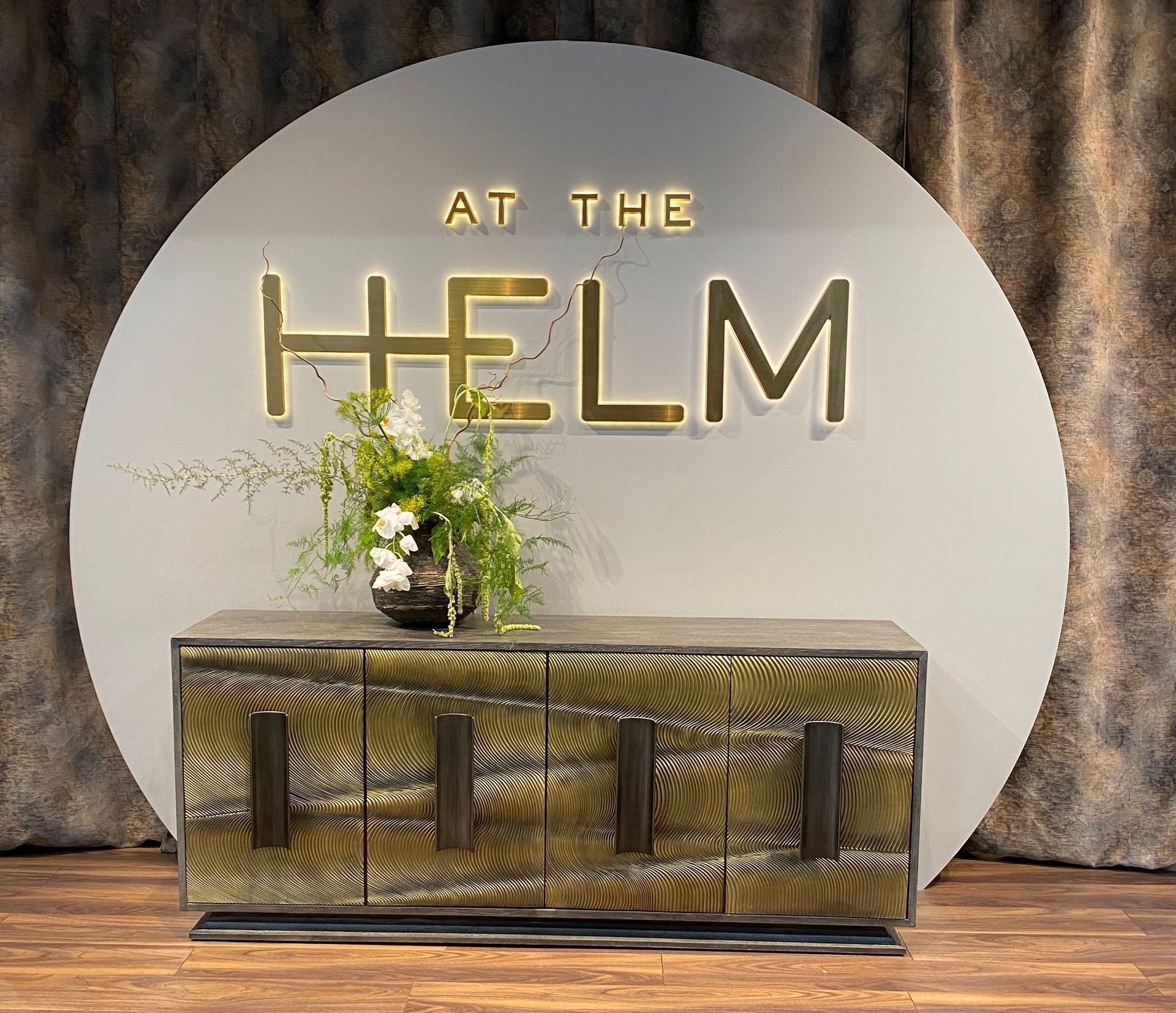 a beautiful luxury 4 door wood cabinet with large gloss wood handles in an abstract brushed gloss metal design with a large floral display sitting on top of the cabinet made by 'At the Helm' seen at the January Furniture Show 2024, NEC, Birmingham