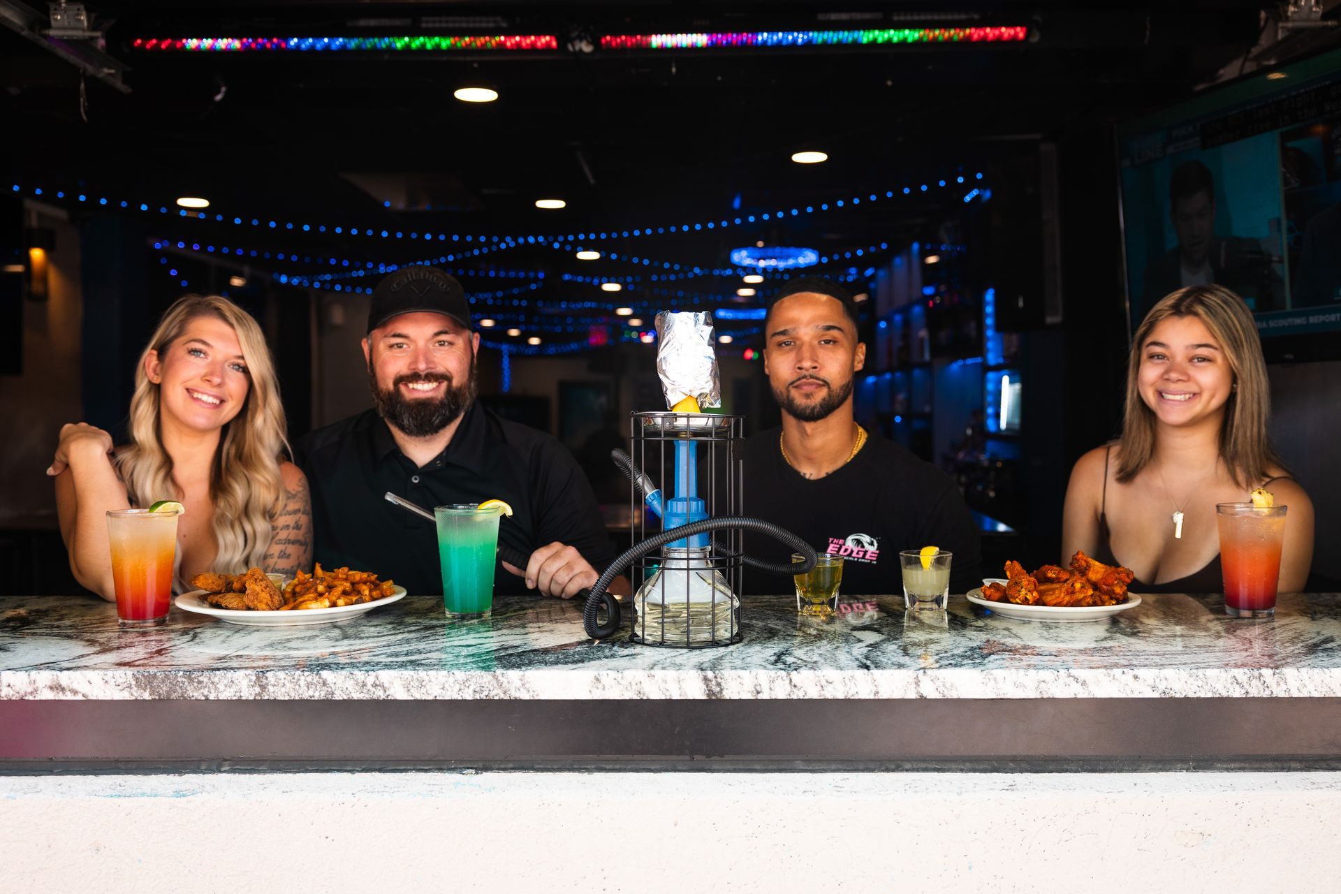 A group of people are sitting at a bar with drinks and food.