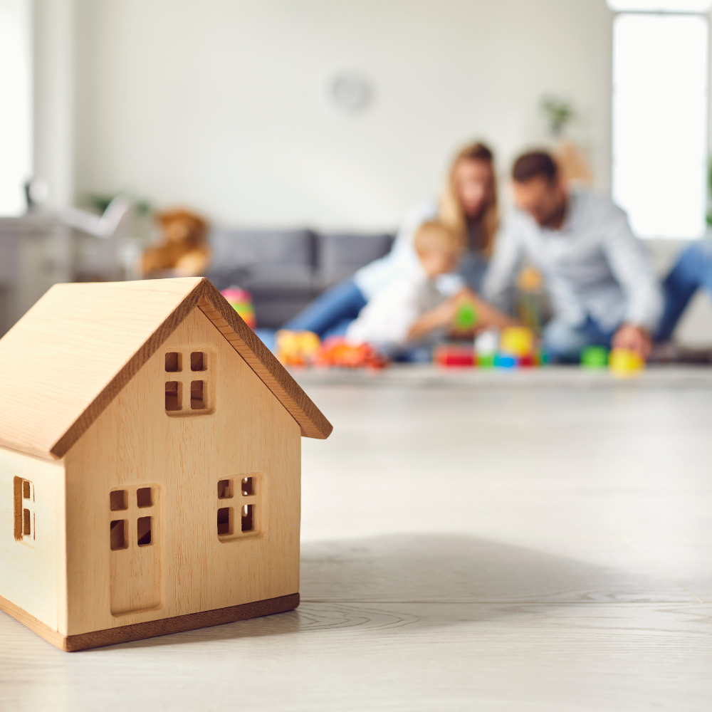 A small wooden house is sitting on the floor in front of a family playing with blocks.