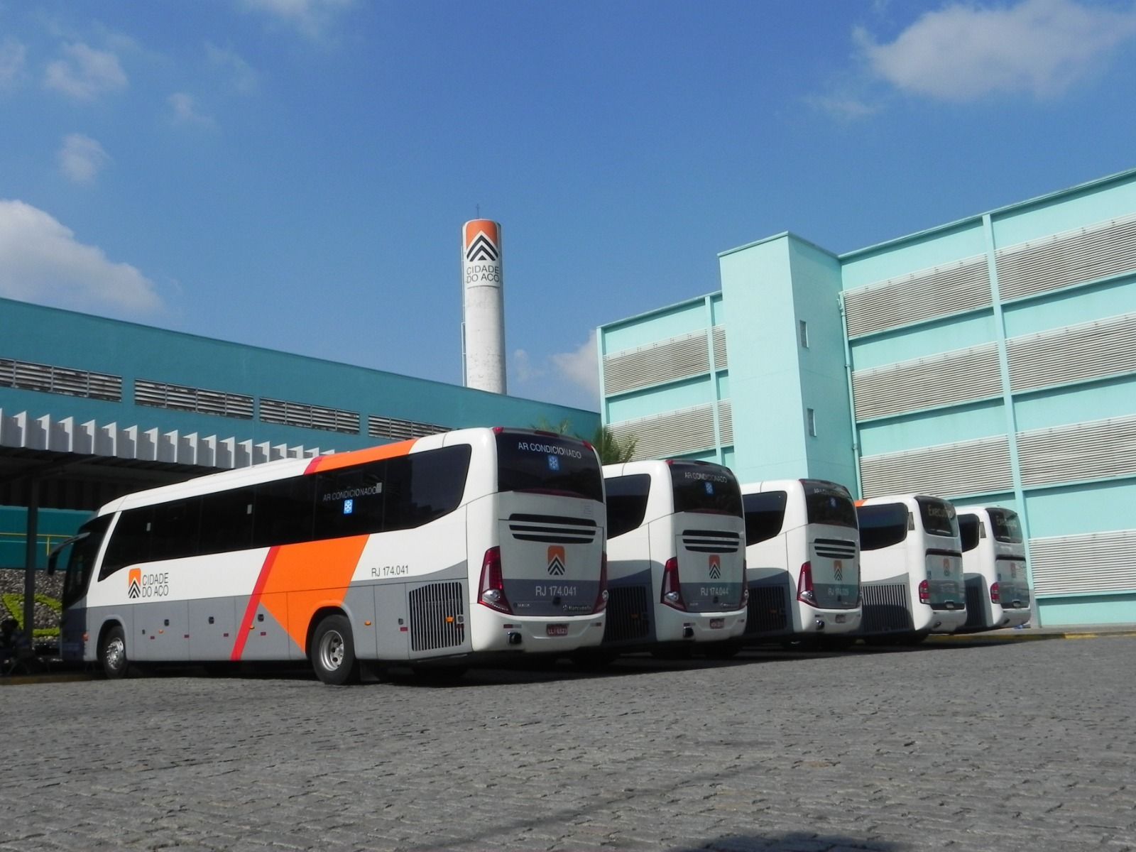 Vários ônibus brancos com detalhes em laranja estacionados em frente a um prédio moderno sob um céu azul.