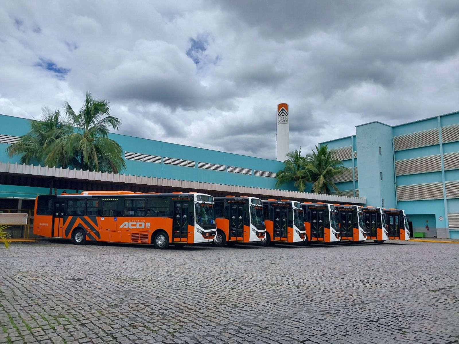 Ônibus laranja estacionados em uma garagem ao lado de um prédio verde-azulado sob um céu nublado.