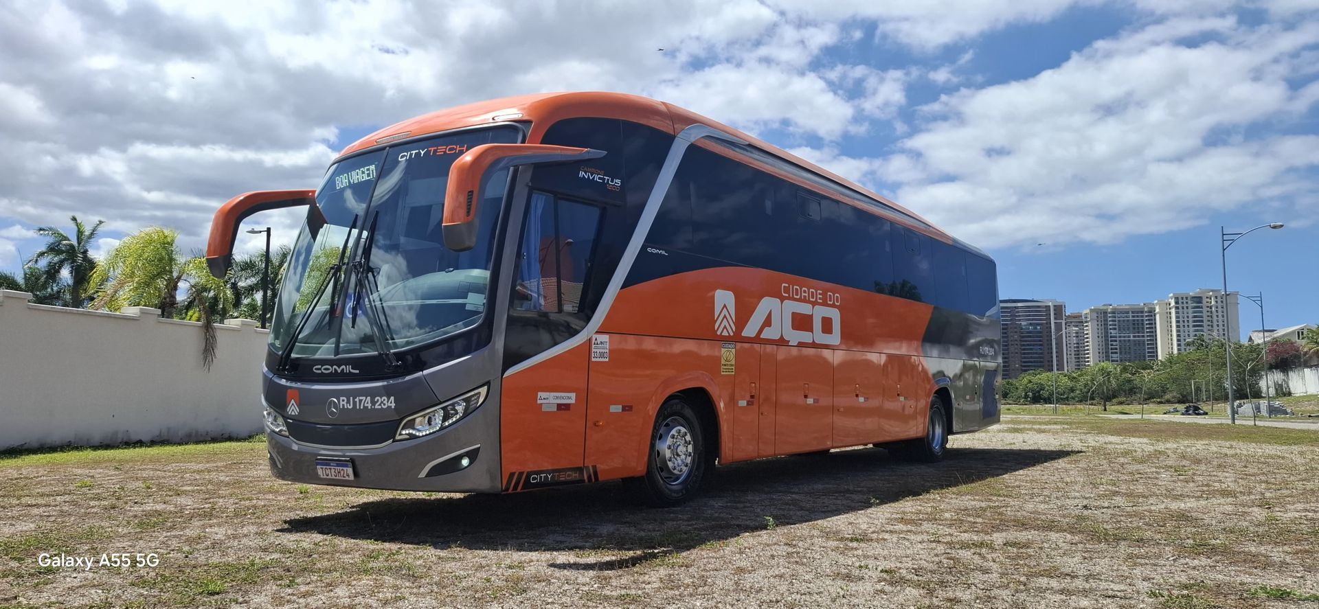 Ônibus de turismo laranja e cinza estacionado em cascalho sob um céu azul.