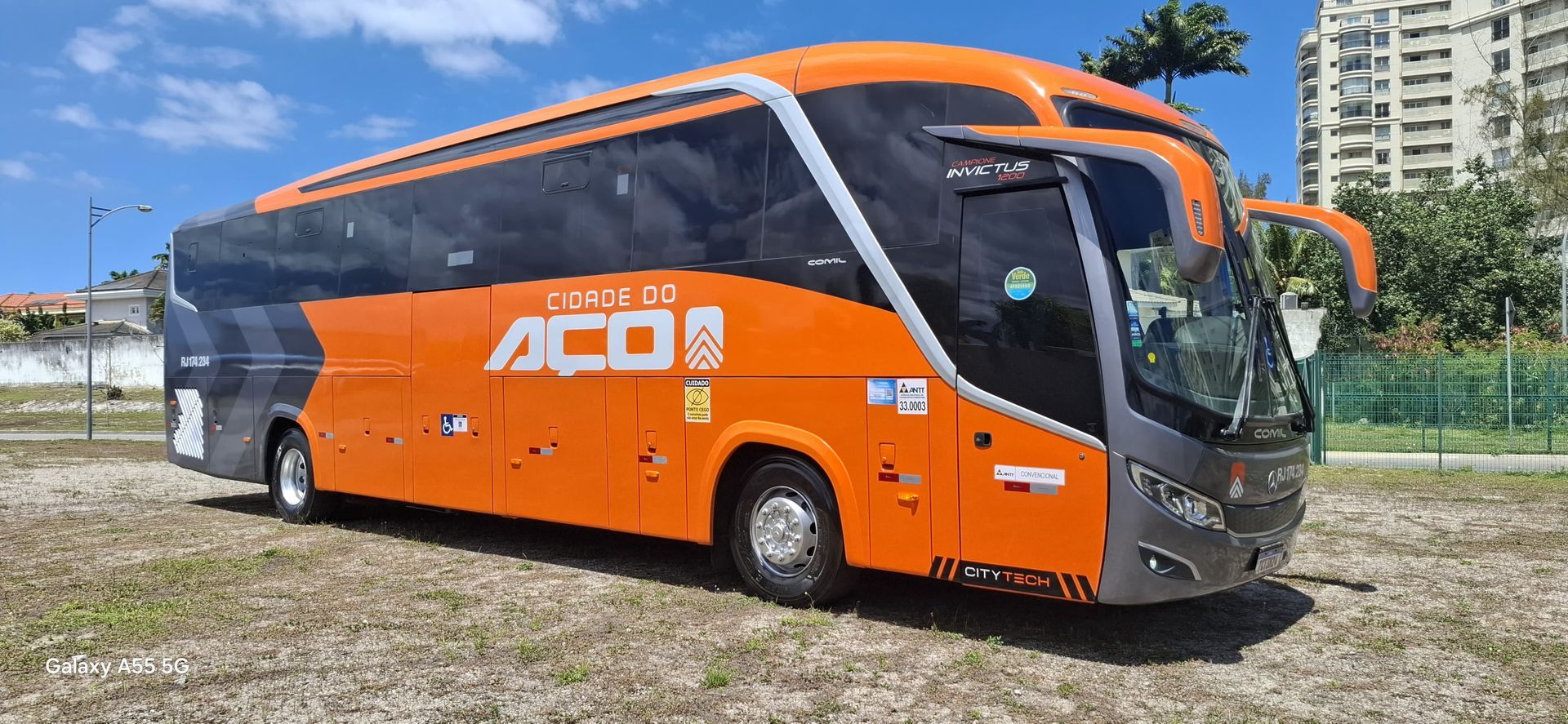 Ônibus laranja da ADO estacionado na grama ao lado de um prédio sob um céu azul.