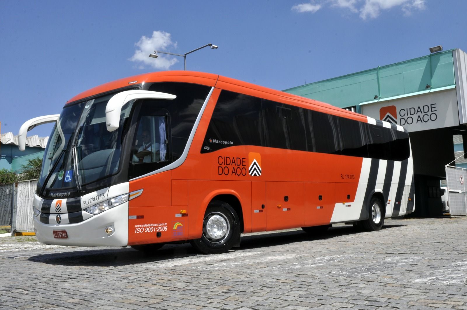 Ônibus turístico laranja e branco estacionado em frente a uma garagem sob um céu claro.