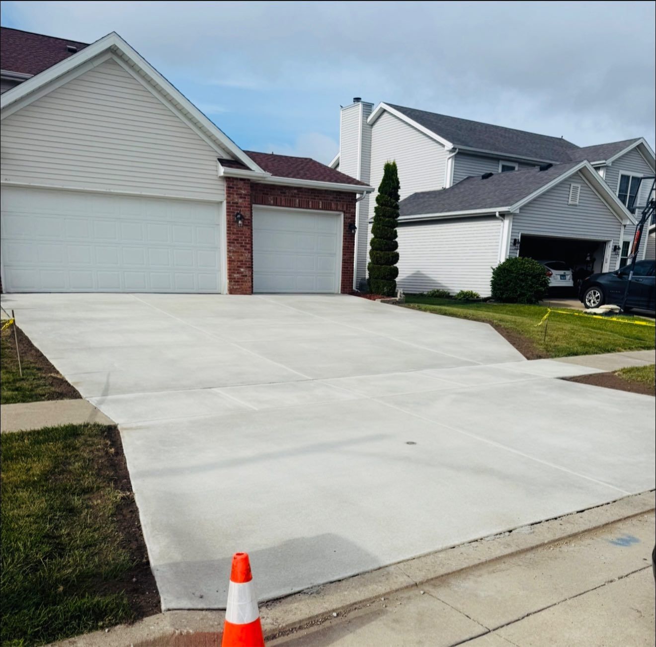A newly poured concrete driveway leads to a two-car garage of a suburban home, with a safety cone in the foreground.