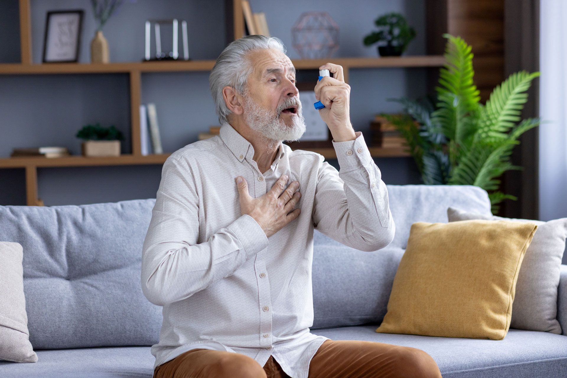 Man using an inhaler, sitting on a couch. He holds chest and looks up; living room background.
