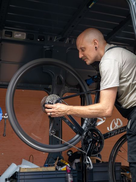 Man repairs a bike wheel inside a van. He’s bald, focused, wearing a t-shirt and overalls.
