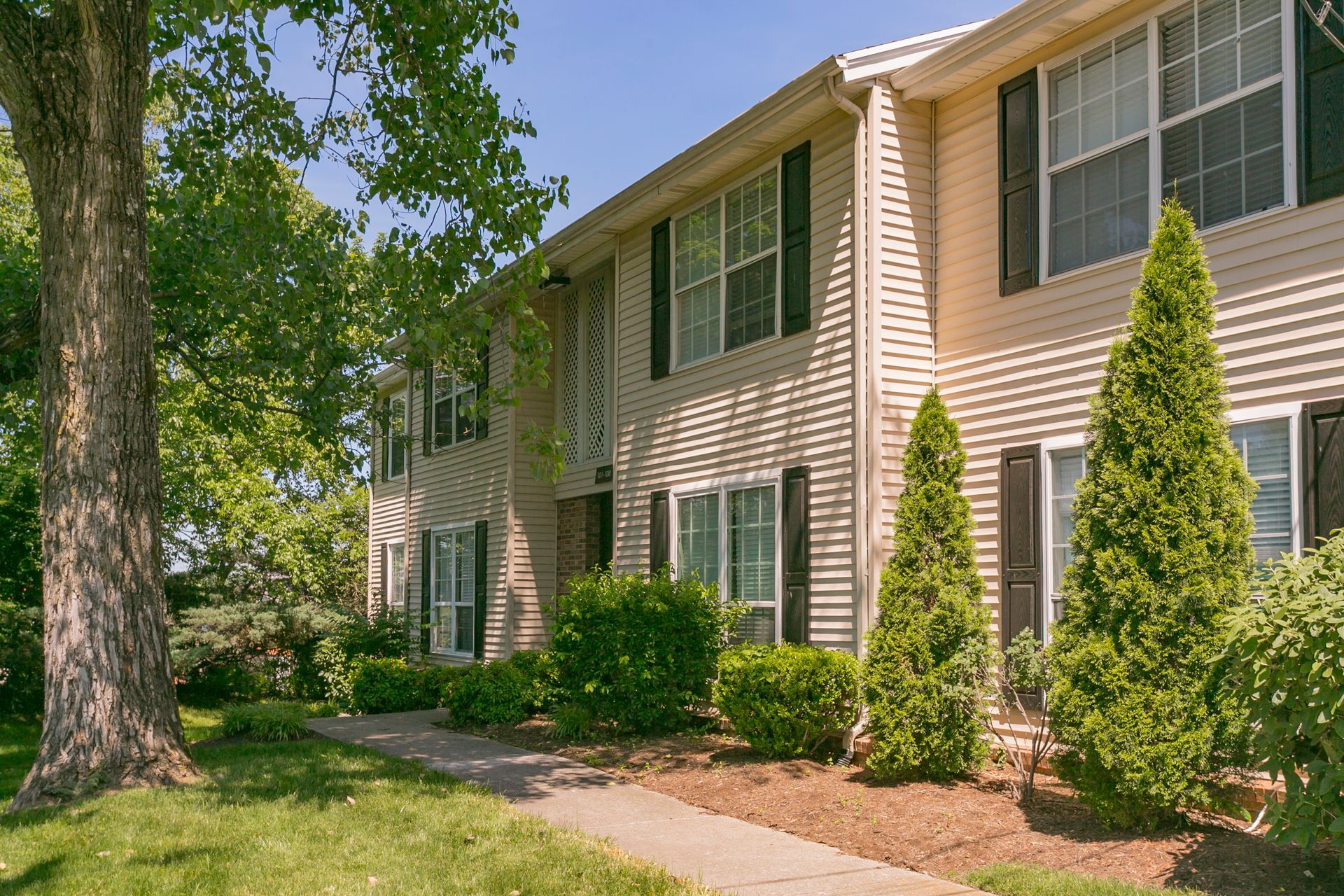 Two-story apartment building with beige vinyl siding and dark shutters, surrounded by green trees and landscaping.