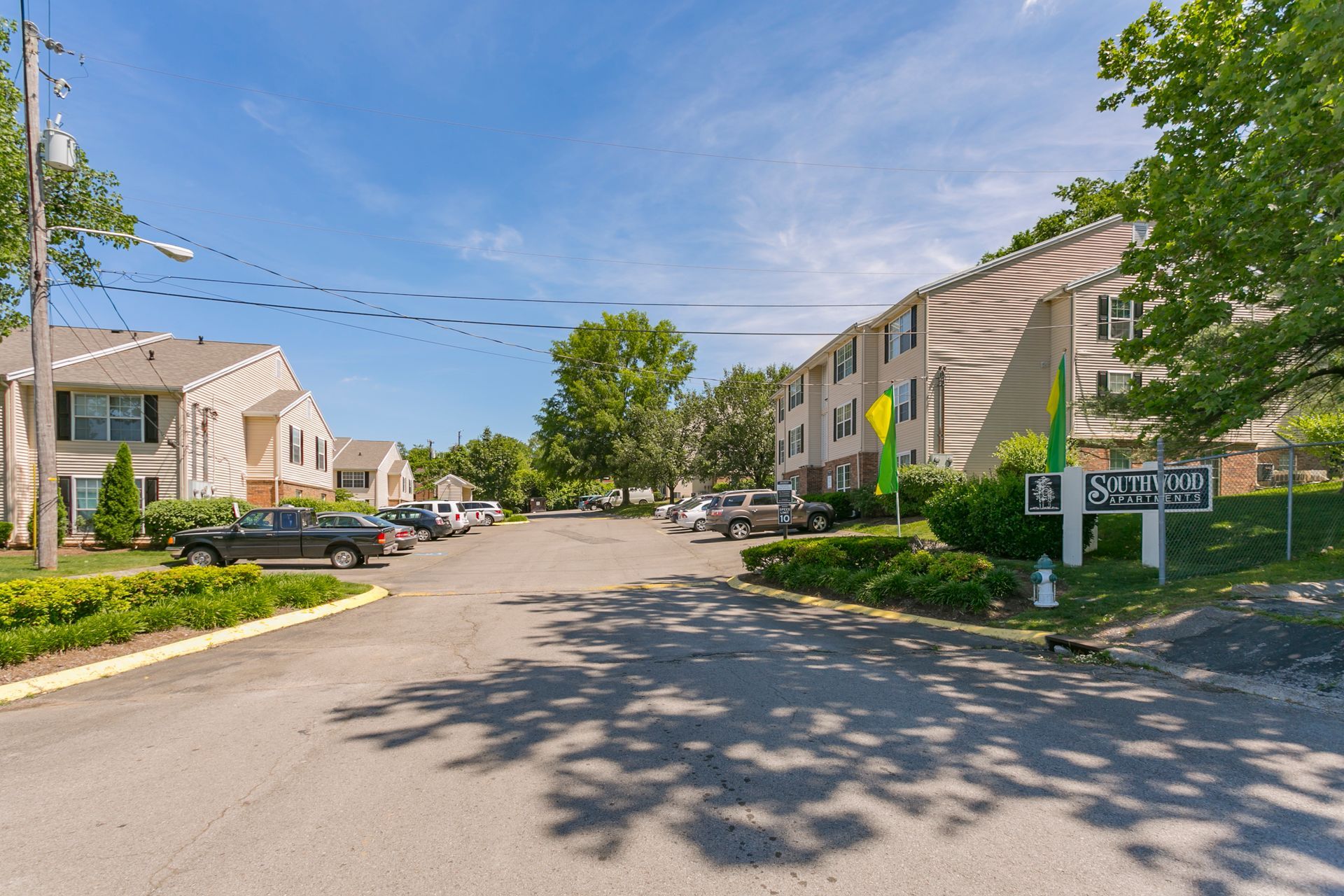 An apartment complex entrance with a paved road, multi-story buildings, parking spaces, and green trees under a blue sky.