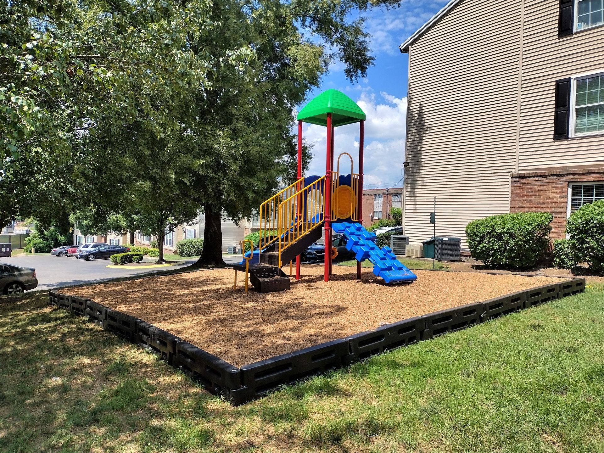 A small playground with a red and green play structure, blue slide, and wood-chip ground, set beside a brick apartment.