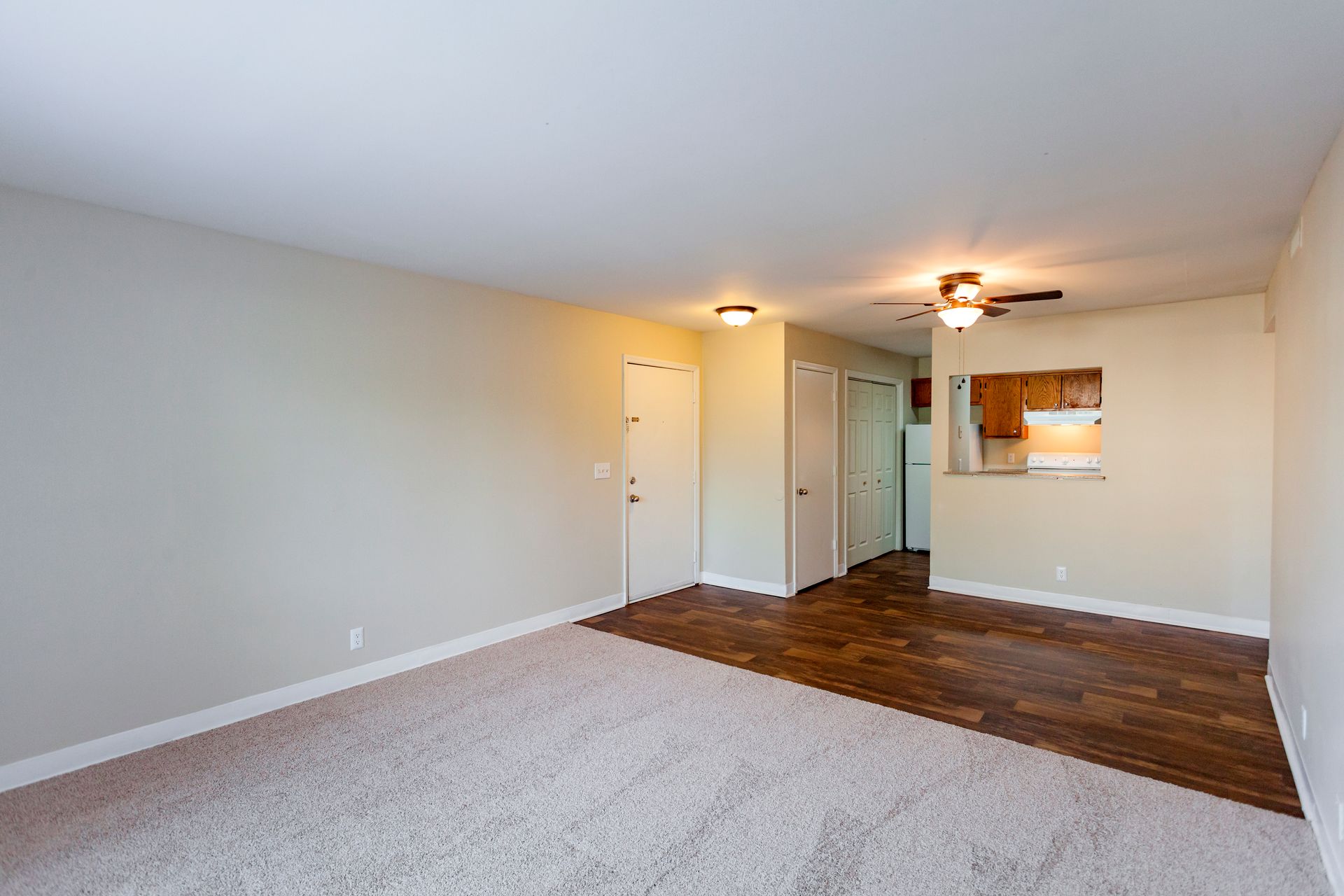 An unfurnished room featuring a beige carpeted living area, wood-look flooring near the entrance, and a kitchen pass-through.