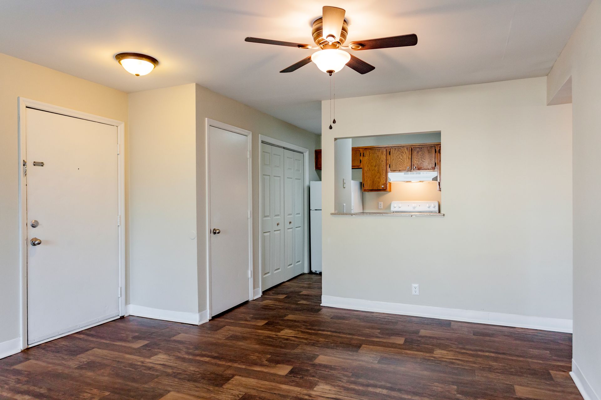 An empty apartment living area with wood-look flooring, cream-colored walls, a ceiling fan, and a kitchen pass-through.