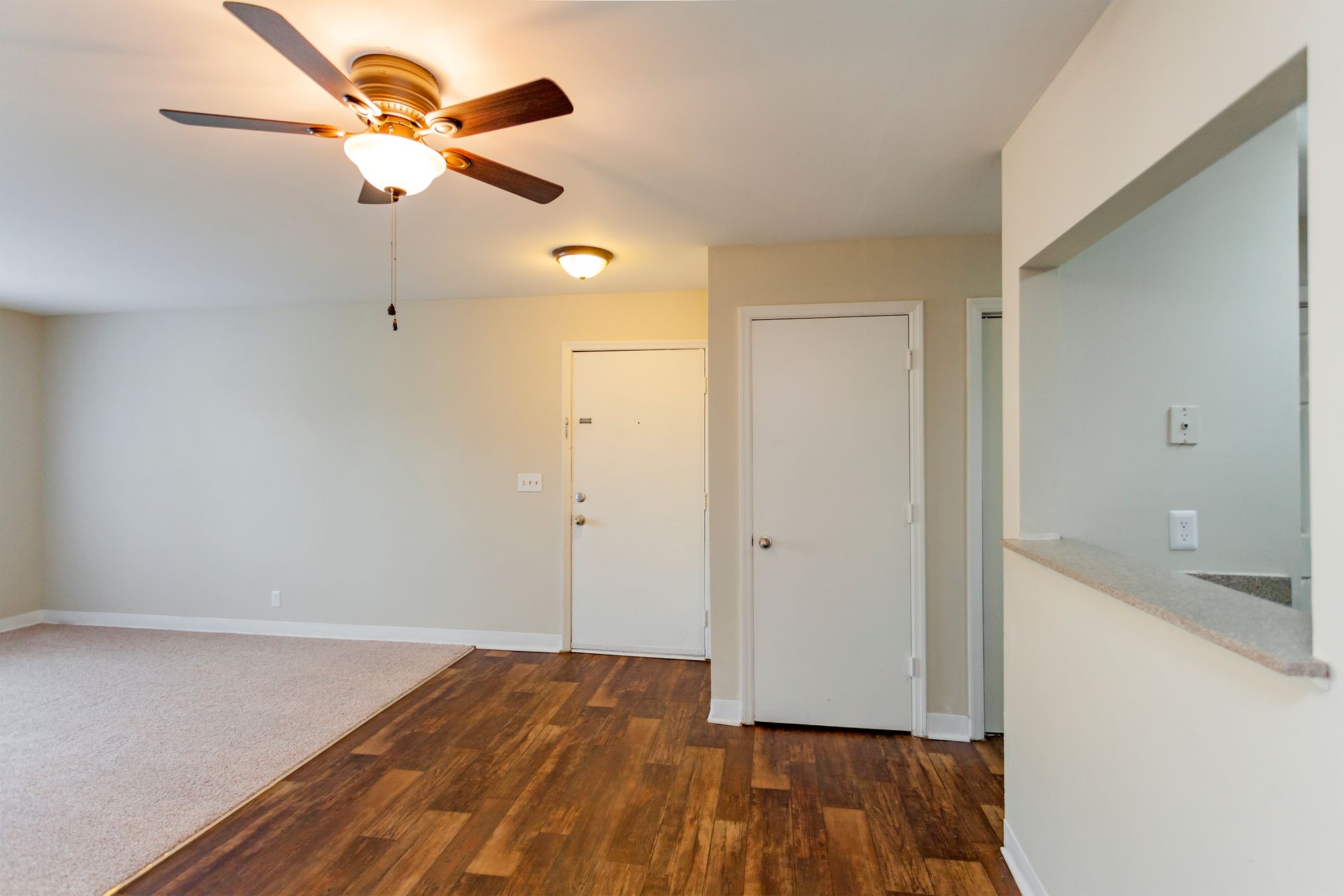 An apartment living room with a ceiling fan, beige walls, carpeted floor, wood-look flooring, and a kitchen pass-through.