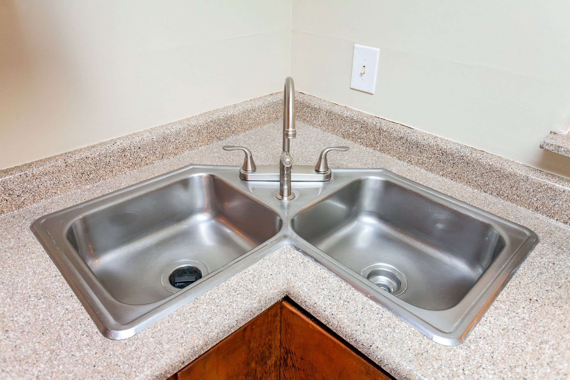 A stainless steel corner sink with two basins and a central faucet set in a light beige speckled countertop.