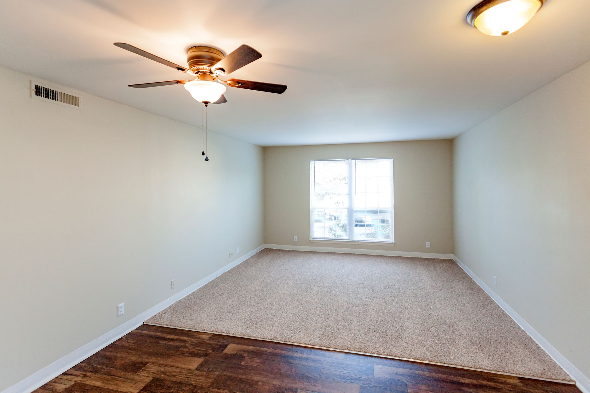 A spacious room with a split floor of dark wood and tan carpet, featuring a ceiling fan and a window at the far end.