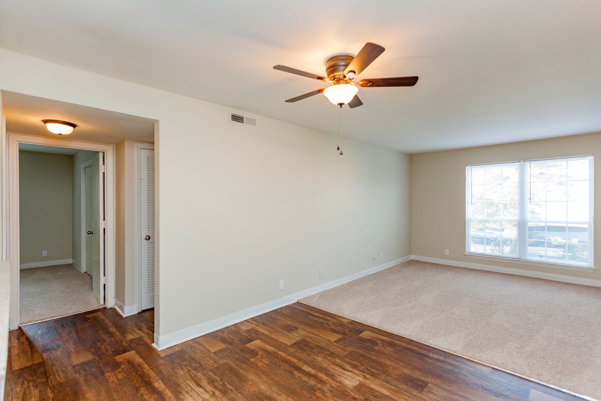 An open interior room featuring wood floors, a carpeted area, a ceiling fan, and a doorway leading to a hallway.