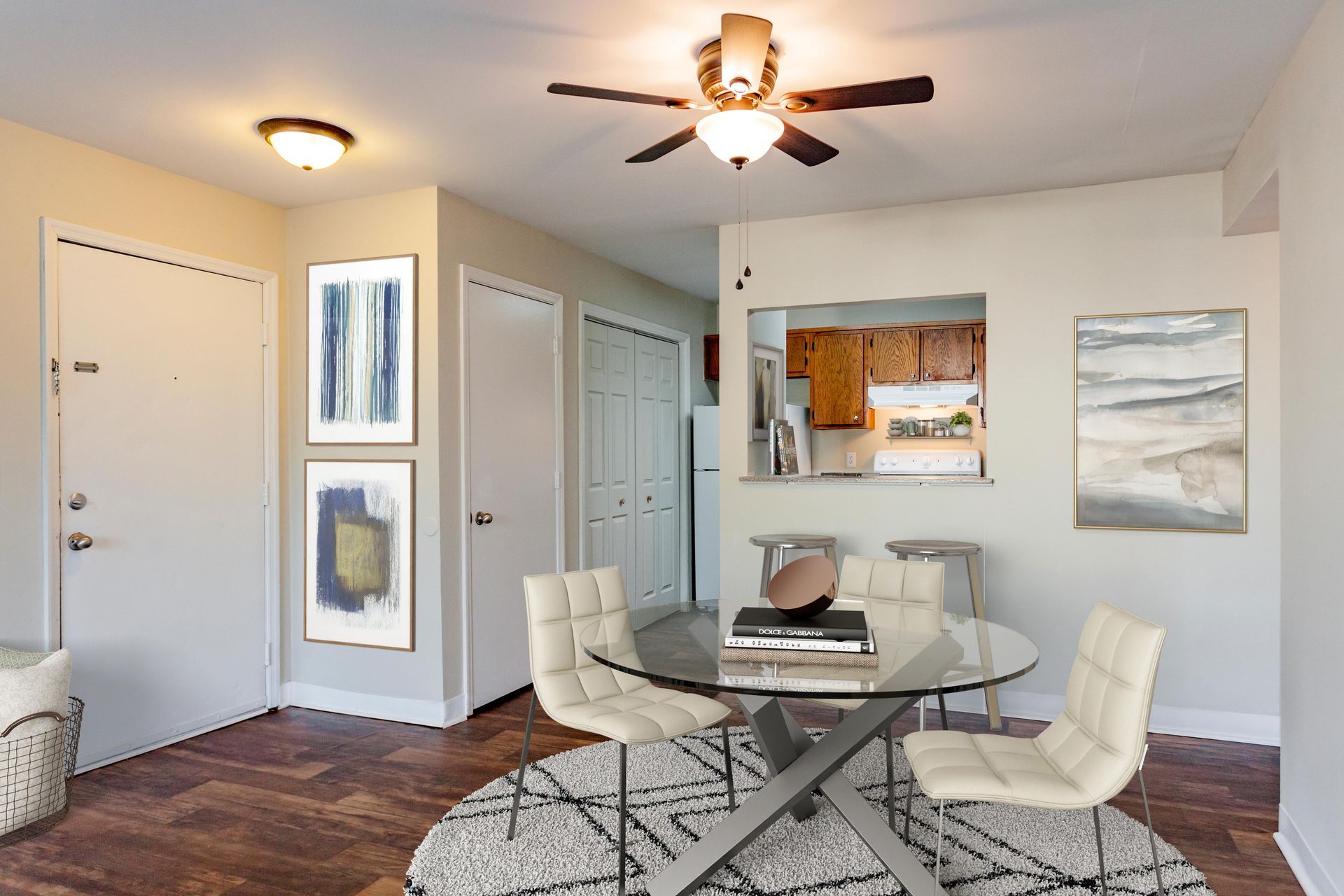 A dining area with a glass table, three white chairs on a patterned rug, two wall art pieces, and a view into a kitchen.