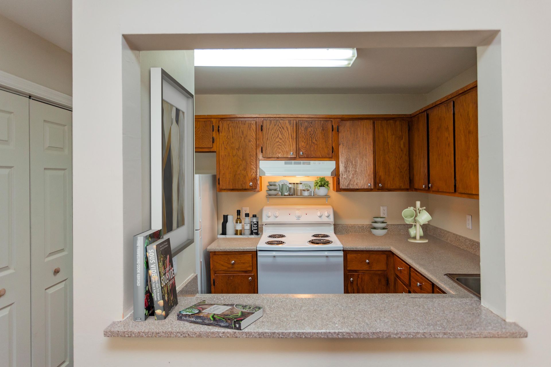 A kitchen viewed through a rectangular opening from an adjacent room, featuring wood cabinets and white appliances.