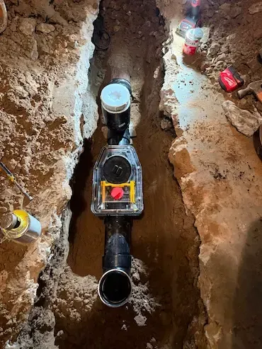 Excavation trench with exposed underground pipes and a valve assembly in muddy soil.