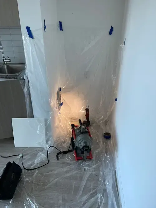 A drain cleaning machine inside a plastic enclosure to contain mess in a room with white walls.