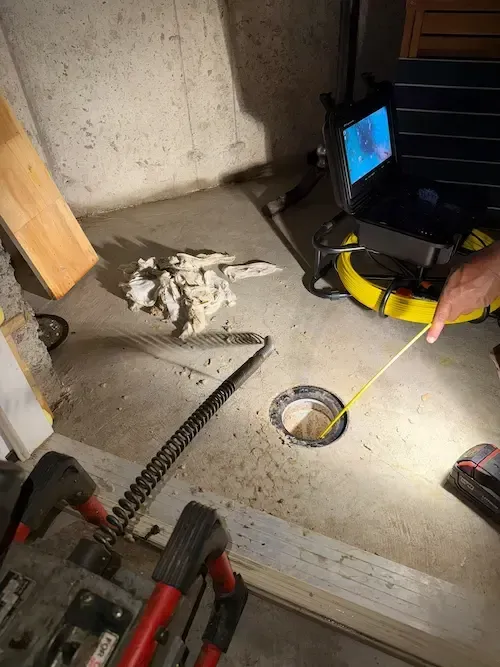 Person measuring a basement floor hole with a yellow tape near tools and a laptop