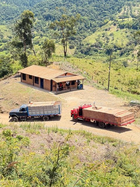 Ladera rural con una pequeña casa y dos camiones que transportan madera en un camino de tierra.