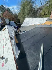 Roofer working on a sloped roof with black shingles and white underlayment near trees and blue sky