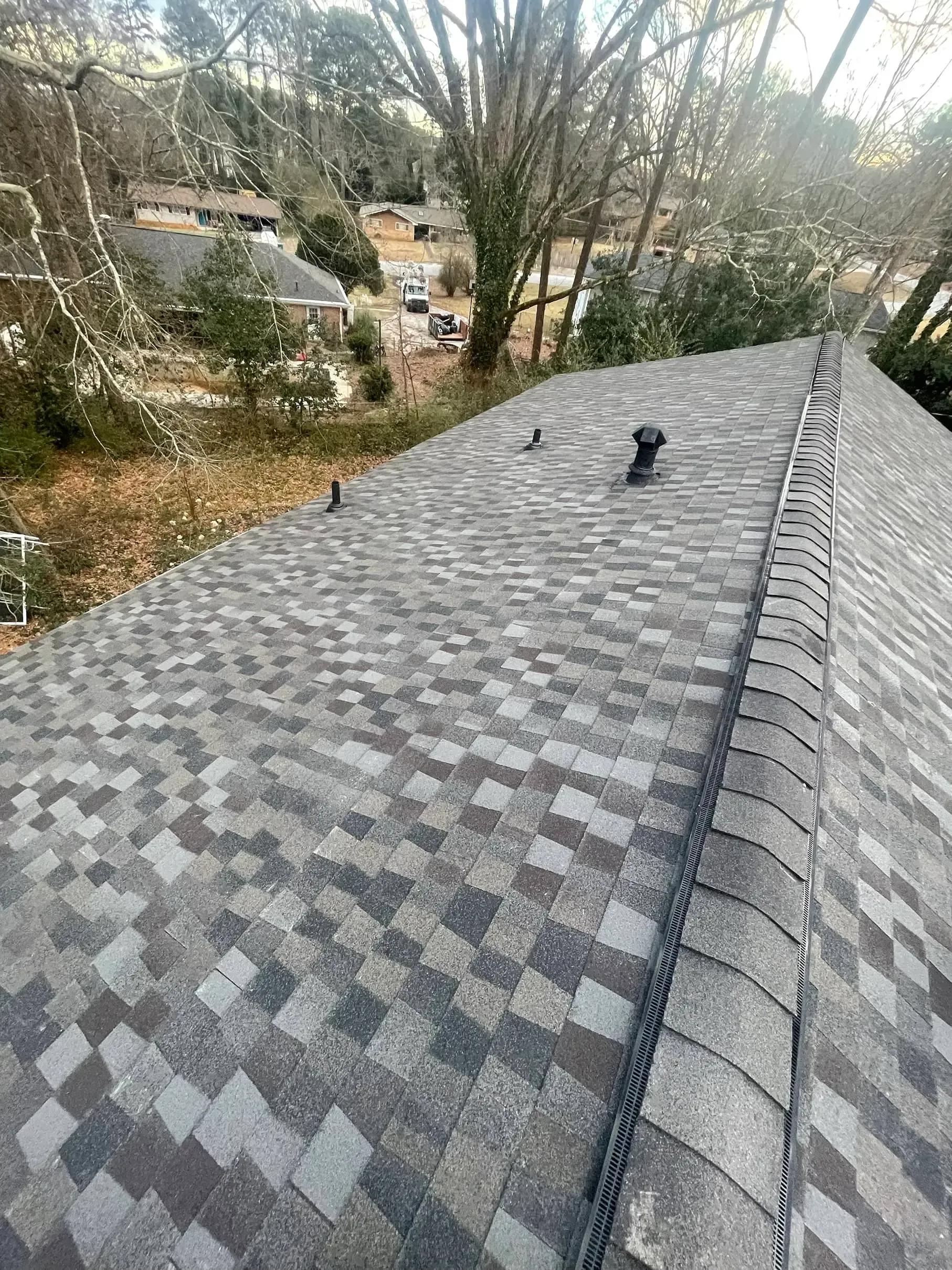 Shingled roof with a vent, overlooking wooded houses in the background.