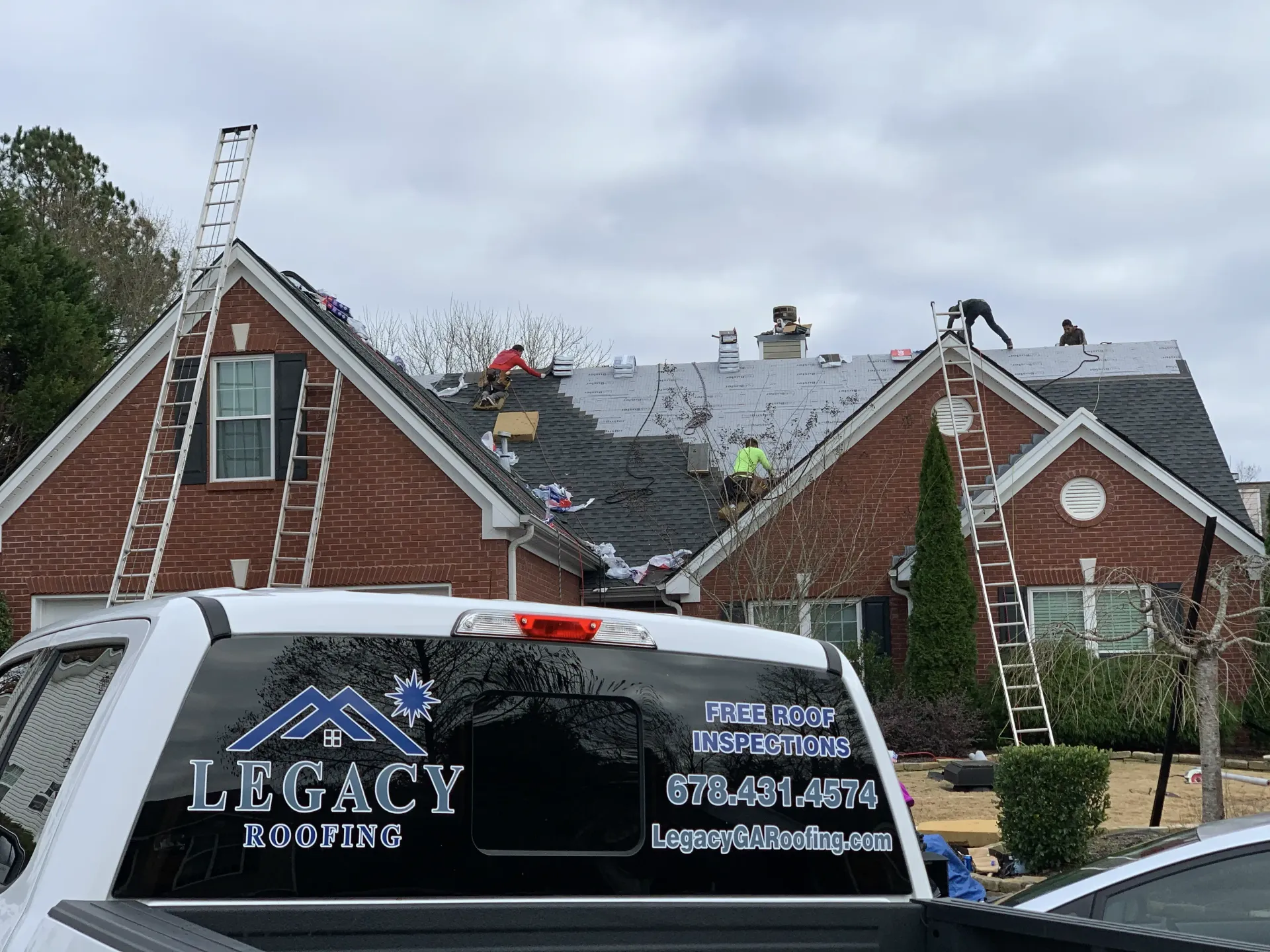 Brick house with roof damage and a roofing company van in front, ladder and workers visible