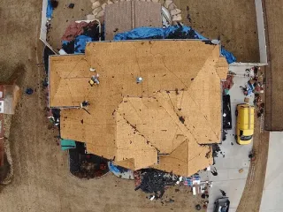 Aerial view of a tan-roofed house with blue tarps and a yellow vehicle beside a curved dirt road.