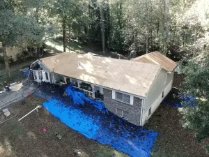 Aerial view of a white house with a tan roof, surrounded by trees and blue tarp covering the yard.