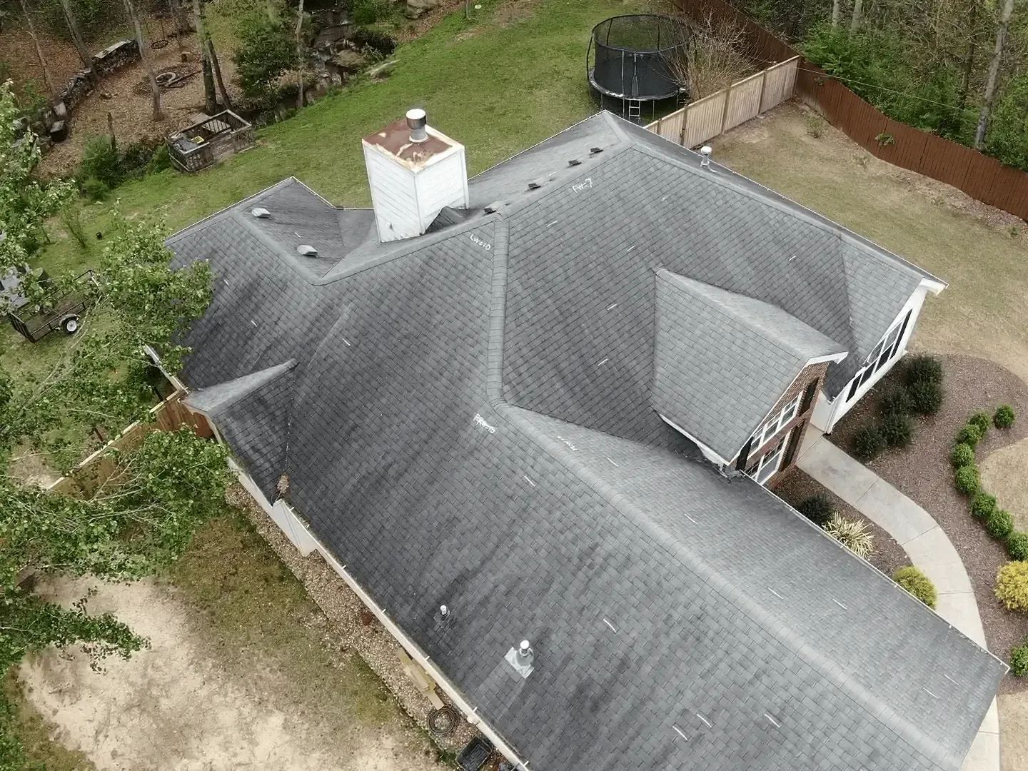 Aerial view of a gray-roofed house with a chimney, surrounded by a yard and shrubs.