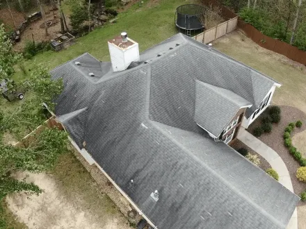 Aerial view of a gray-roofed house with a chimney, surrounded by a yard and shrubs.