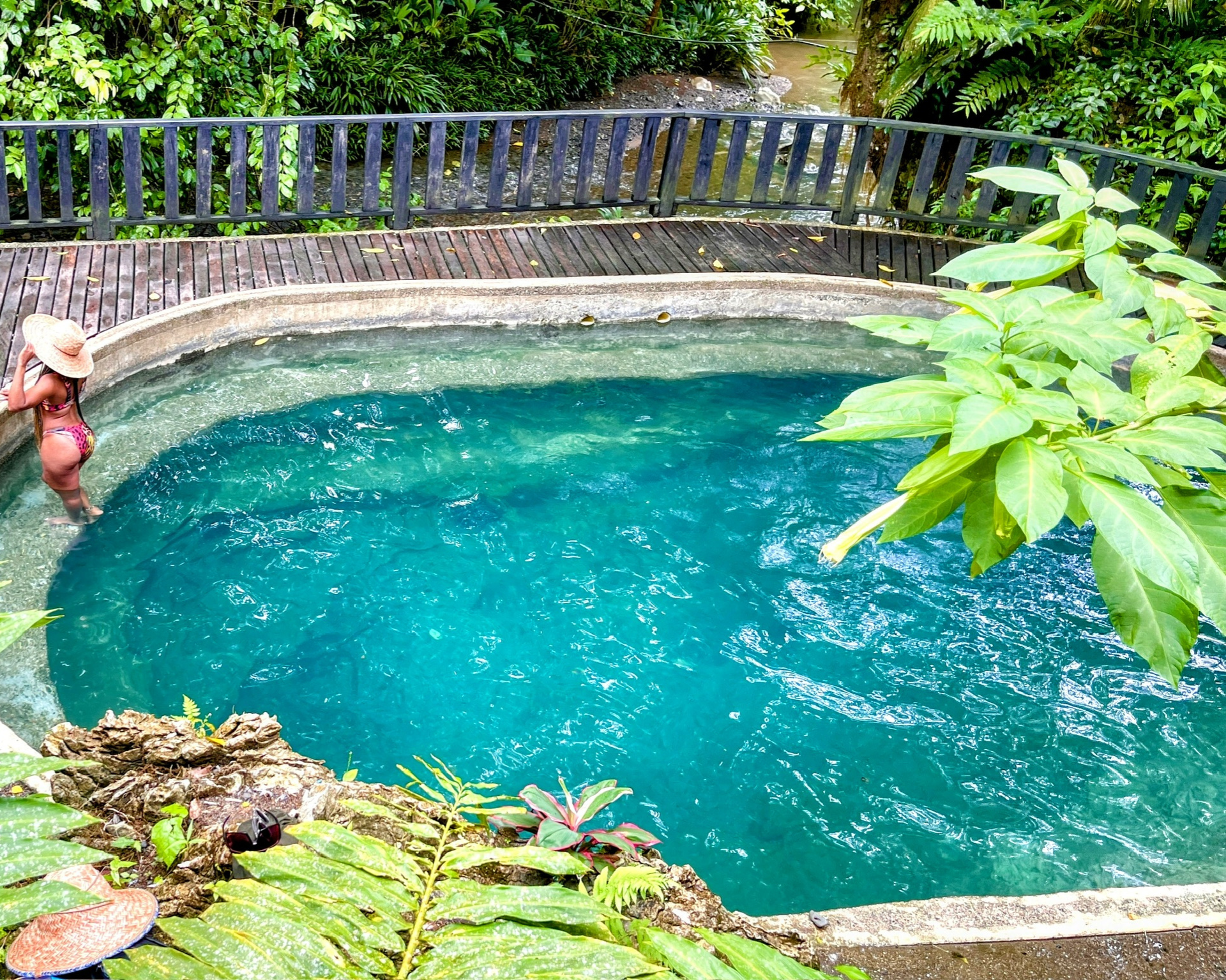 Una mujer en traje de baño se encuentra en una piscina azul claro rodeada de exuberante vegetación y una valla de madera.