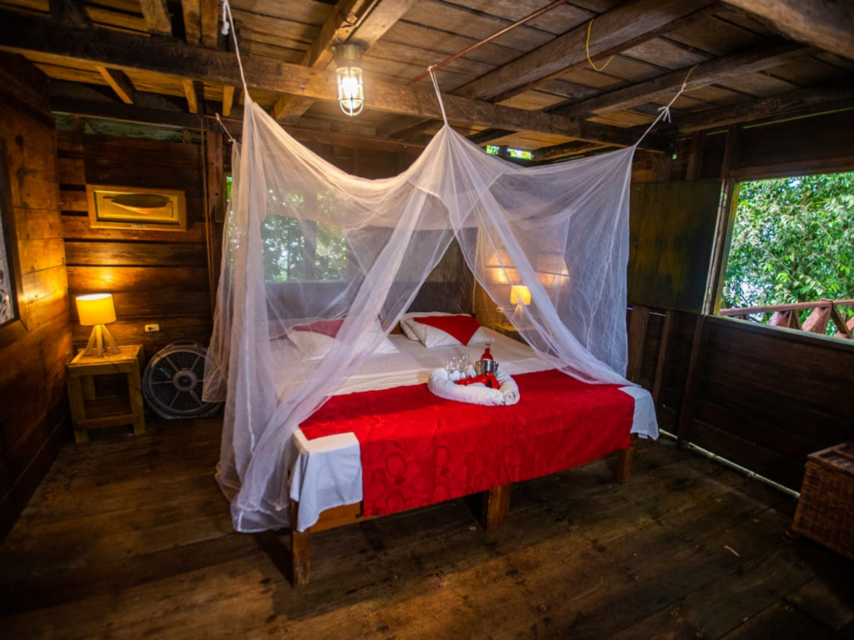 Interior de cabaña de madera con cama, mosquitero, sábanas rojas y ventana con vista a los árboles.