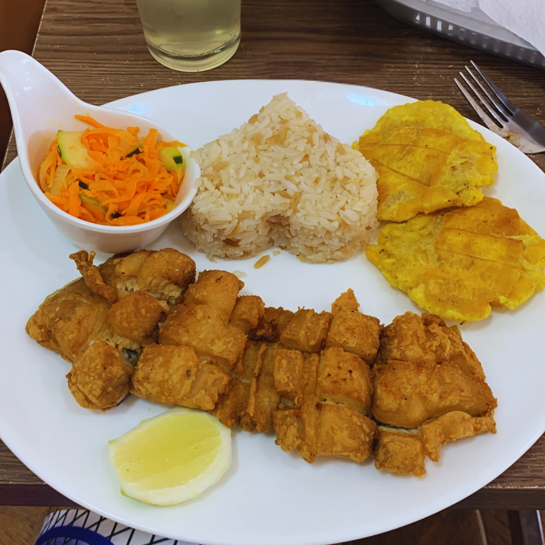 Plato con pescado frito, arroz, plátanos y ensalada. También incluye una rodaja de limón y un vaso de líquido.