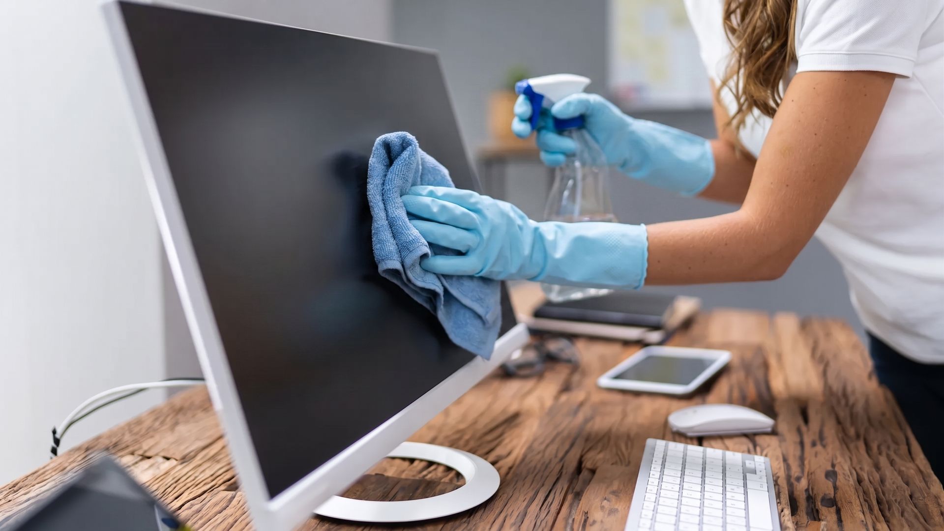 Person cleaning a computer monitor with a blue cloth and spray bottle at a desk