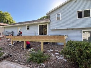 Workers building a wooden deck beside a light gray house in a rocky backyard.