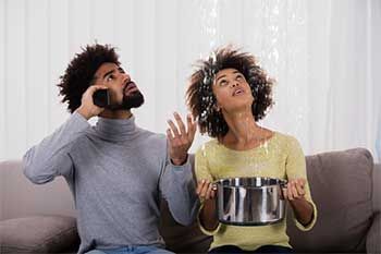 Couple indoors, looking up at a water leak from ceiling; woman holds a pot, man on phone.