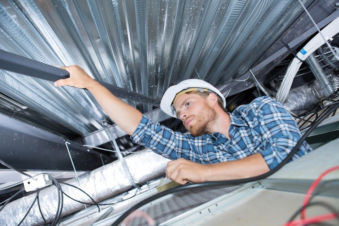 Construction worker inspecting ceiling ductwork, wearing a hard hat, reaching upward.