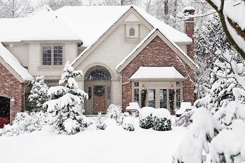 House covered in snow, snowy yard with evergreen trees. Beige siding, red brick, brown door.