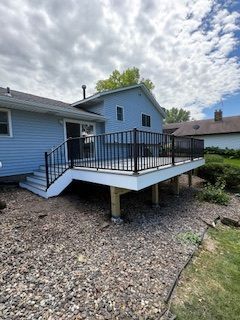 Blue house with elevated deck, black railing, and stairs over a gravel yard under a cloudy sky