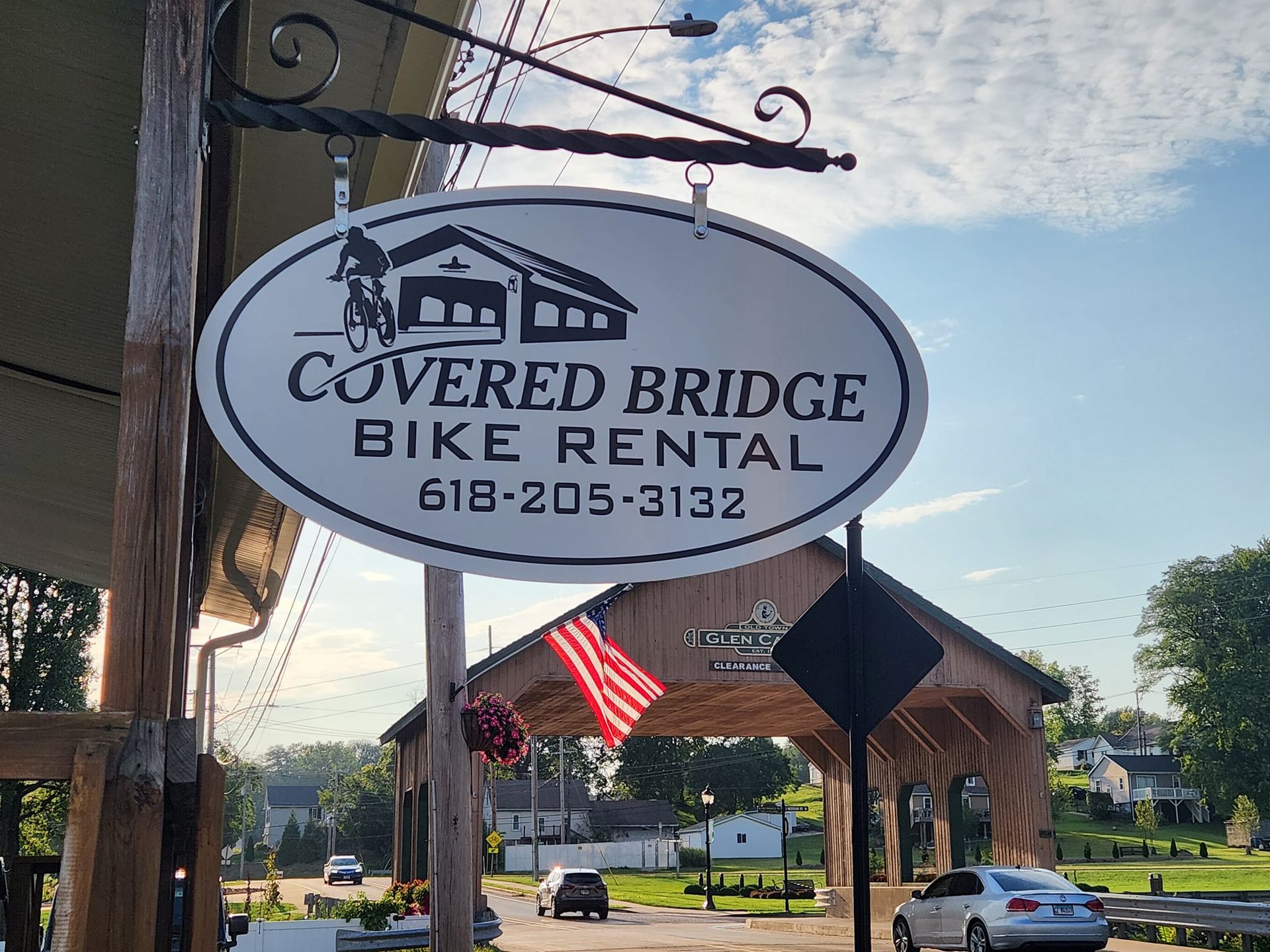 Oval sign for Covered Bridge Bike Rental with phone number, hanging outside a shop with an American flag.