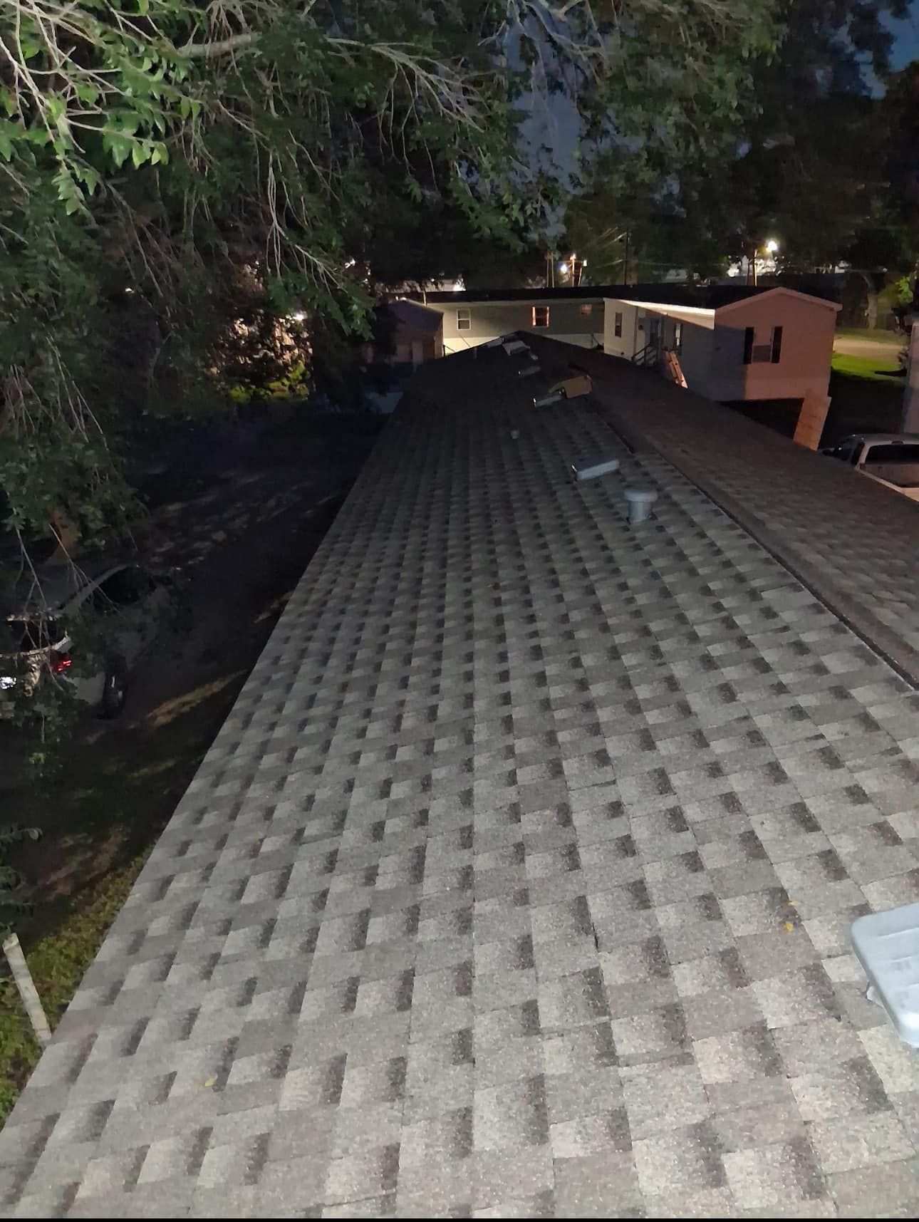 Dark photo of a roof covered in gray shingles, with some trees and buildings in the background.