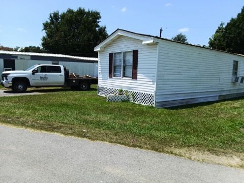 White mobile home with a parked pickup truck on a grassy lot beside a paved road.
